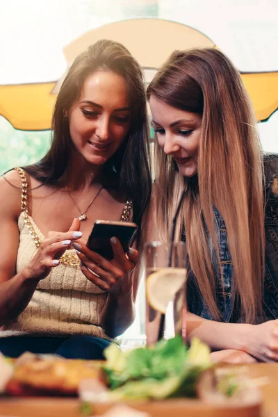 Twee glimlachend Kaukasische vriendinnen kijken naar foto's en video's op smartphone zit in koffie winkel met lunchpauze — Stockfoto