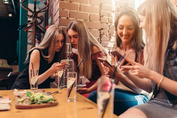 Cuatro estudiantes sonrientes sentadas en la cafetería charlando con teléfonos móviles — Foto de Stock