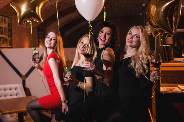 Glimlachend Europese vriendinnen gekleed in Cocktailjurken drinken alcoholhoudende dranken met bachelorette partij in trendy restaurant — Stockfoto