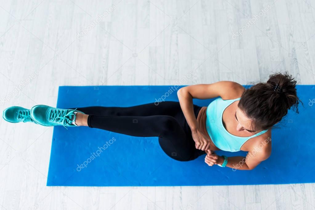 Top view of young fit woman working-out doing bicycle crunches on blue mat indoors.