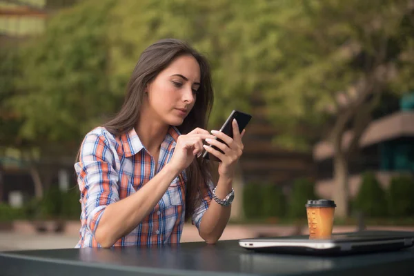 Atractiva joven leyendo un mensaje de texto en su teléfono celular. Chica sentada al aire libre usando smartphone —  Fotos de Stock