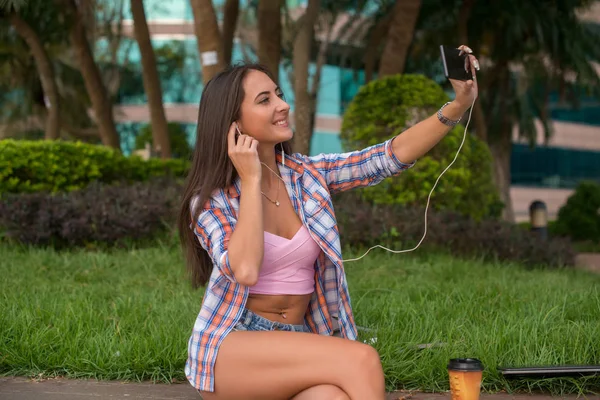 Szczęśliwa młoda kobieta noszenia słuchawek i robienie zdjęć w parku siedzi na krawężniku. Atrakcyjna kobieta selfie. — Zdjęcie stockowe