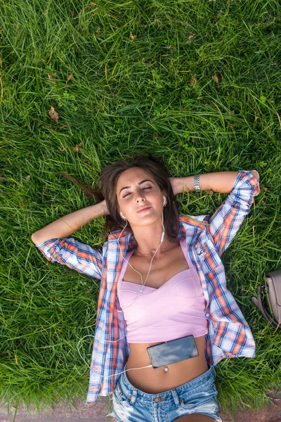 Mujer joven y relajada con auriculares escuchando música desde un teléfono inteligente y tumbada en la hierba con los ojos cerrados —  Fotos de Stock