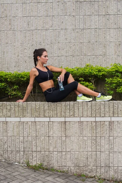 Mujer joven relajándose después de hacer deporte, sentada contra la pared de piedra gris, escuchando música, sosteniendo una botella de agua, preparándose para correr . — Foto de Stock