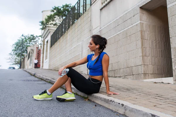 Unga athletic kvinnliga vila efter kör motion, lyssna på musik i hörlurar, håller en flaska vatten, sitter på trottoaren gata. — Stockfoto