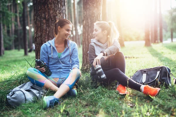 Two young women in sportswear sitting under trees in the forest drinking water, talking and resting after training outdoors — Stock Photo, Image