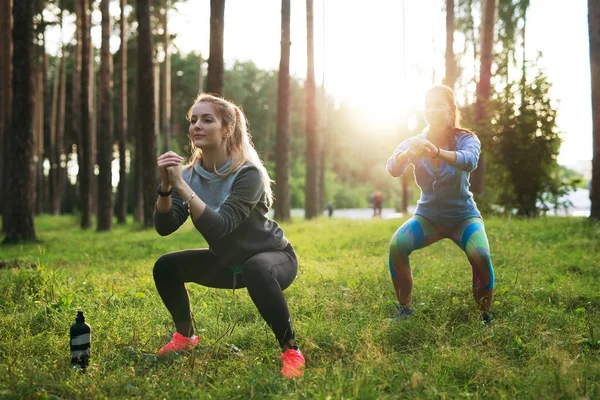 Fitte junge kaukasische Frauen in Jumpsuits, die frühmorgens im Wald Kniebeugen machen — Stockfoto