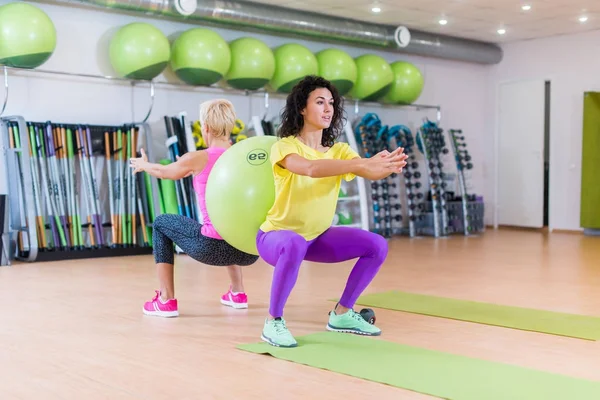 Zwei junge Frauen, die Kniebeuge-Übungen machen, mit einem schweizerischen Ball dazwischen. Sportlerinnen trainieren im Fitnessstudio. — Stockfoto