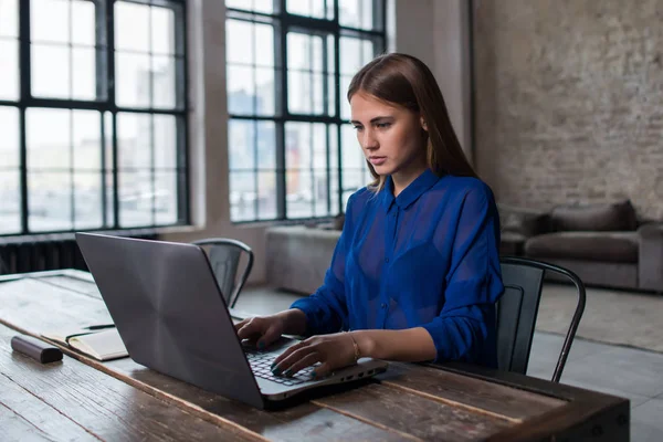 Ganska ung brunett arbetar på laptop på träbord i rymliga mörka loft studio — Stockfoto