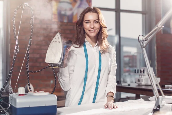 Retrato de la atractiva joven modista morena caucásica de pie en ropa blanca sosteniendo plancha de vapor en el estudio . — Foto de Stock