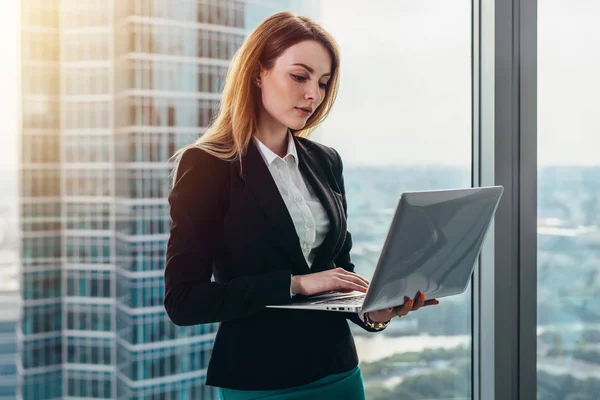 Mladé samice právník pracující v luxusní kanceláře drží notebook stojící proti panoramatická okna s výhledem na obchodní čtvrti — Stock fotografie