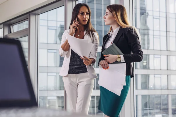 Deux femmes d'affaires détenant des documents parlant à leur client par téléphone mobile offrant différentes solutions debout dans le bureau à la mode — Photo