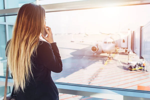 Młoda kobieta interesu rozmowy na telefon komórkowy, czekając na jej lotu, patrząc na samoloty przez okno stojące w strefie wyjazdu na lotnisko — Zdjęcie stockowe