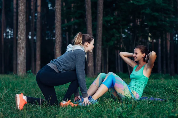 Sonriente chica delgada haciendo abdominales mientras amiga ayudándola a sostener los pies. Dos novias entrenando juntas en el bosque . —  Fotos de Stock