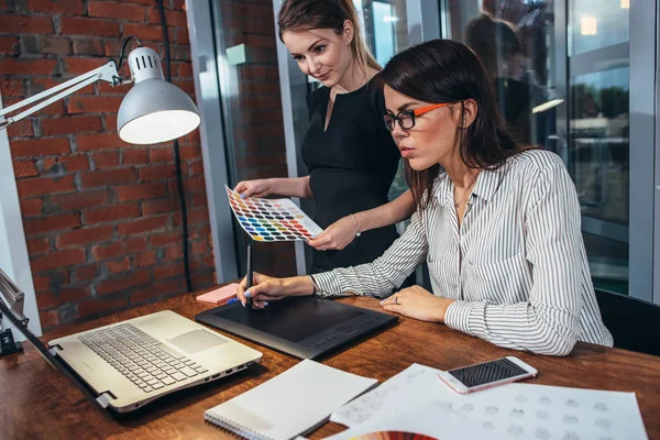 Team av kvinnliga inredningsarkitekt rita ett nytt projekt med hjälp av grafisk tablett, laptop och färgpalett som sitter vid skrivbord i moderna studio — Stockfoto