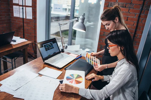 Jovens mulheres trabalhando em um novo web design usando amostras de cores e esboços sentados na mesa no escritório moderno — Fotografia de Stock