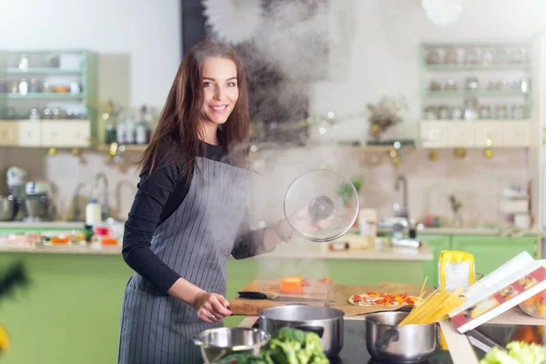 Mujer bastante joven con delantal haciendo la cena cocinando espaguetis siguiendo la receta en un libro de pie en la cocina —  Fotos de Stock