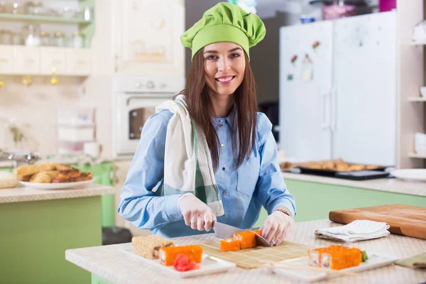 Cocinera con sombrero de chef y guantes haciendo rollos de sushi japonés, sonriendo, mirando a la cámara en la cocina — Foto de Stock