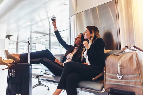 Duas mulheres conversando selfie enquanto esperam por um voo de atraso sentado com malas no terminal do aeroporto — Fotografia de Stock