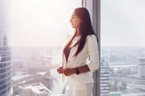 Retrato de uma jovem chefe pensativa e bem-sucedida que está perto da janela no escritório olhando para a paisagem urbana — Fotografia de Stock