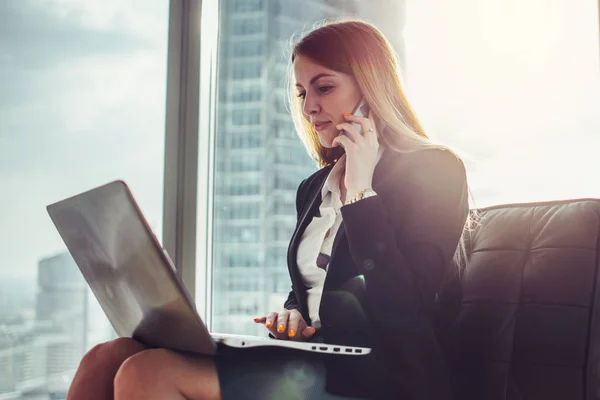 Jovem esperando em um salão sentado no escritório moderno trabalhando no laptop falando ao telefone — Fotografia de Stock