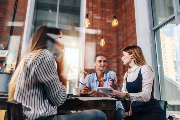 Equipo de tres jóvenes empresarias pensando en un nuevo proyecto de negocio anotando las ideas sentadas a la mesa en oficce — Foto de Stock