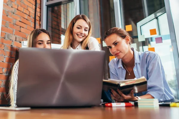 Три юные девушки разговаривают онлайн-уроком с помощью ноутбука, сидящего в библиотеке — стоковое фото