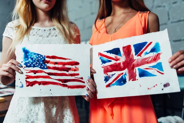 Close-up image of two young women holding a drawing of British and American flags hand-drawn with aquarelle technique on plain paper — Stock Photo, Image