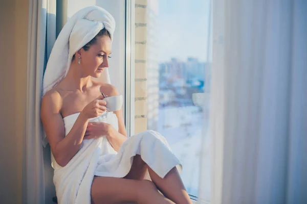 Romantische Frau sitzt vor dem Fenster und bewundert Sonnenaufgang oder Sonnenuntergang mit Handtuch auf dem Kopf Körper nach dem Bad. — Stockfoto