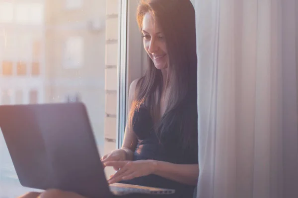 Kvinnan ler och använder bärbar dator hemma sitter nära fönster — Stockfoto