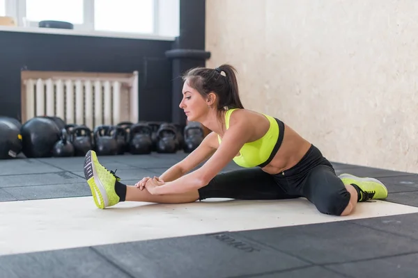 Mujer joven estirando isquiotibial en el gimnasio, en forma femenina haciendo calentamiento en la estera — Foto de Stock