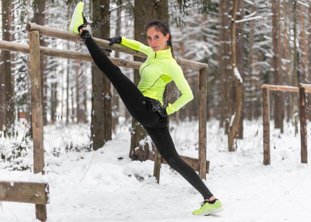 Fit woman athlete doing left leg split stretching exercises outdoors in woods. Female sports model exercising outdoor winter park