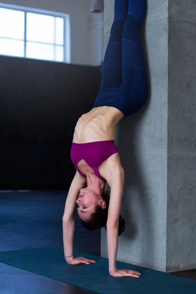 Slender young woman standing in inverted yoga pose performing handstand near the wall in sports studio — Stock Photo, Image