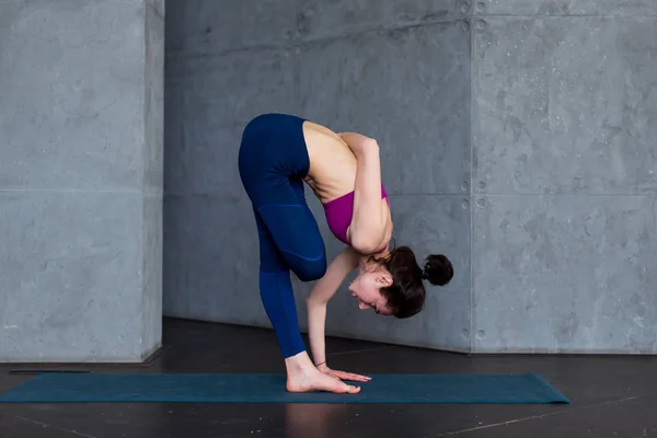 Flexible young woman performing one-leg balance exercise doing half bound lotus standing forward fold pose indoors