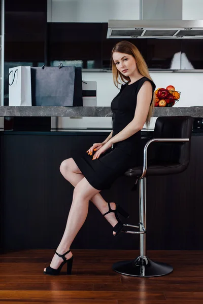Full-length portrait of elegant woman with fair hair wearing black dress and high heels sitting on a bar chair in kitchen