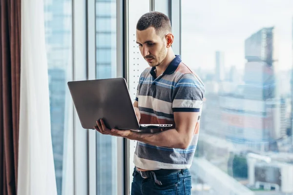 Affärsman som håller en laptop arbeta stående i kontor nära fönstret med utsikt över staden — Stockfoto
