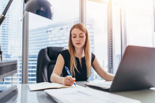 Chefe executiva feminina sentada em sua mesa tomando notas em agenda escrevendo com caneta e usando seu computador no moderno prédio de escritórios — Fotografia de Stock