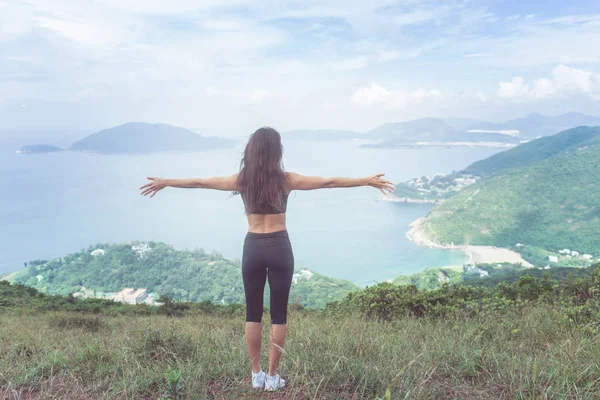 Achteraanzicht van fitness vrouw stond op groene berg met haar uitgestrekte armen kijken naar zee landschap uiting van geluk en vrijheid — Stockfoto