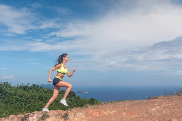 Mujer atlética corriendo por la montaña con el cielo y el mar de fondo. Corredor profesional haciendo ejercicio cardiovascular al aire libre en el paisaje natural —  Fotos de Stock