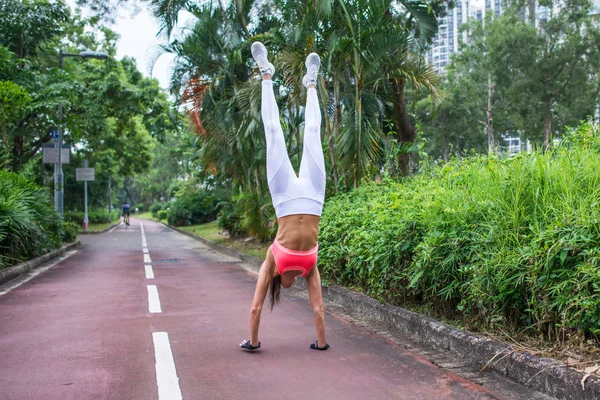 Vista frontal de una joven en forma delgada que practica yoga haciendo parada de mano recta en el día de verano del sendero del parque. Chica deportiva haciendo ejercicio al aire libre . —  Fotos de Stock