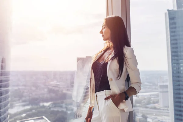 Ritratto di elegante donna d'affari che indossa abito formale bianco in piedi vicino alla finestra guardando paesaggio urbano — Foto Stock