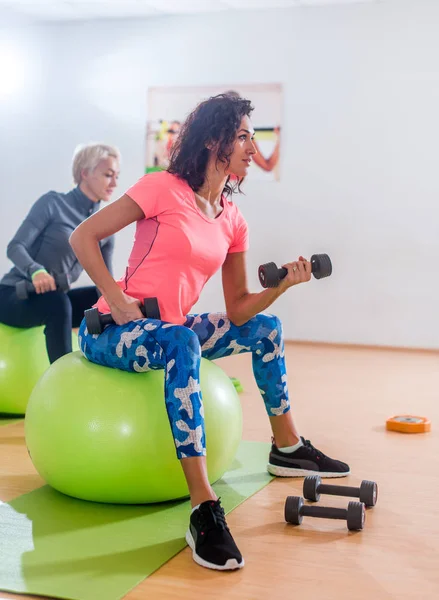 Donne magre sportive che partecipano alla lezione di fitness in palestra che si allenano sedute su palle fisiche facendo bicipiti alternati arricciati con manubri — Foto Stock