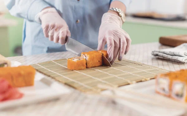 Imagem cortada de mulher em luvas cortando rolos de sushi em esteira de bambu e servindo pratos — Fotografia de Stock