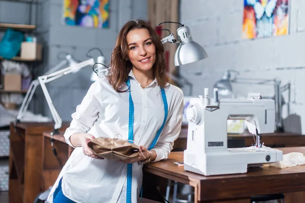 Portrait of smiling European fashion designer standing next to sewing machine holding a gift packed in craft paper in studio — Stock Photo, Image