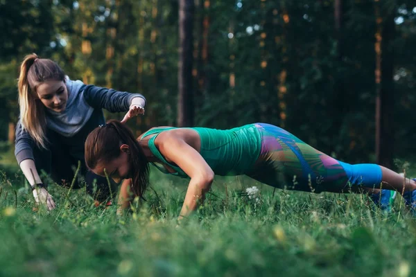 Инструктор по фитнесу помогает молодой женщине делать отжимания на траве в парке — стоковое фото