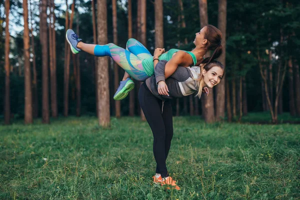 Молодая улыбающаяся женщина поднимает свою подругу на спину, развлекаясь во время тренировки в лесу — стоковое фото
