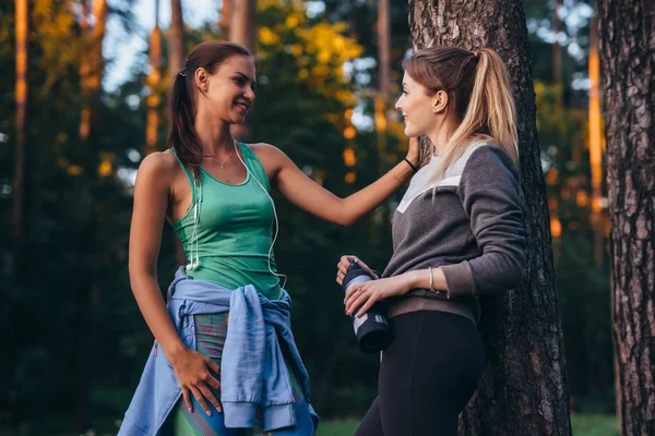 Duas corredoras relaxando após o treino em pé perto da árvore falando no parque — Fotografia de Stock