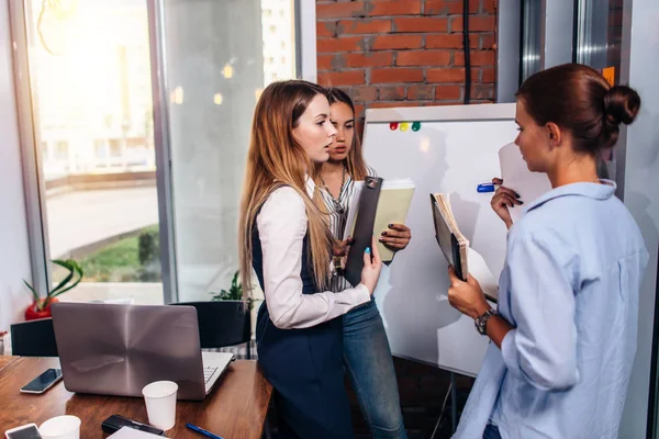 Tre giovani imprenditrici che condividono nuove idee imprenditoriali in possesso di note in piedi alla lavagna bianca in carica — Foto Stock