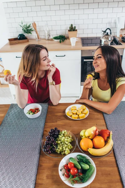 Deux amies assises dans la cuisine, mangeant des fruits et légumes . — Photo