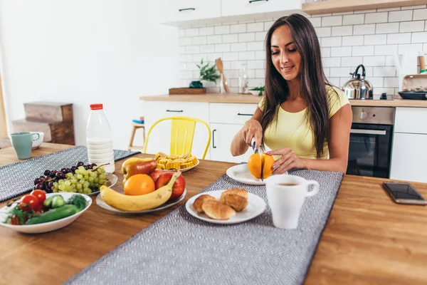 フルーツ、オレンジ、コーヒー、クロワッサンで朝食します。台所のテーブルに座っている女性 — ストック写真